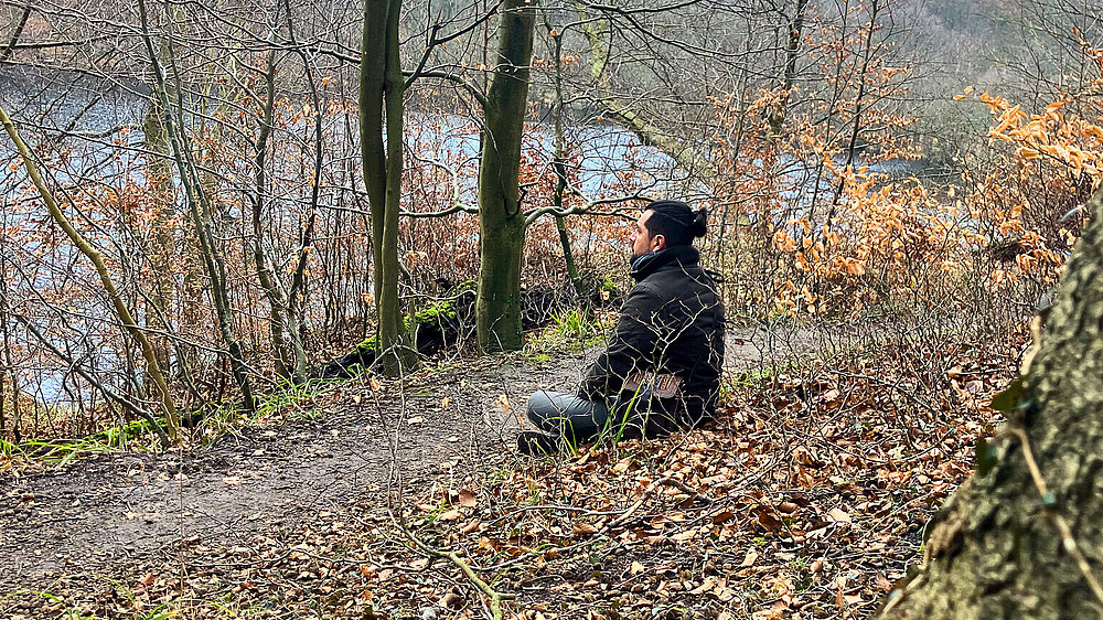 Ein Mann sitzt am Waldrand auf einem Weg und blickt auf einen stillen See, umgeben von kahlen Bäumen und vereinzeltem, braunem Laub. Die Umgebung vermittelt eine ruhige, natürliche Atmosphäre.