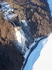 Bild - Luftaufnahme einer winterlichen Landschaft mit einem Kreidefelsen, umgeben von Bäumen, die teilweise schneebedeckt sind. Am Fuße des Felsens erstreckt sich ein gefrorener See. Im Hintergrund sind Strukturen und Wege sichtbar.