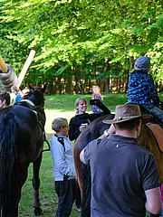 Bild - Eine Gruppe von Menschen versammelt sich im Freien, umgeben von Bäumen und Pferden. Ein Junge sitzt auf einem Pferd, während Erwachsene miteinander sprechen und Freude teilen. Einige halten Getränke in der Hand, die Stimmung ist entspannt und gesellig.