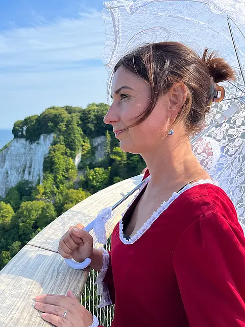 Eine Frau in einem roten Kleid steht auf einer Aussichtsplattform, hält einen weißen Stockschirm und blickt auf das Meer und die steilen Klippen im Hintergrund. Der Himmel ist blau mit wenigen Wolken, und die Umgebung ist grün bewaldet.