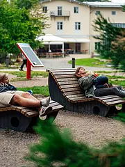 Bild - Eine Familie entspannt auf zwei modernen Liegen im Freien. Ein Erwachsener und zwei Kinder liegen bequem, genießen die Ruhe und die Natur um sie herum. Im Hintergrund sind Bäume und eine Spielplatzumgebung zu sehen.