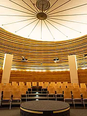 Bild - Ein moderner Konferenzraum mit einer runden Holzdecke. In der Mitte steht eine kreisförmige Bühne, umgeben von halbkreisförmig angeordneten Holzstühlen. Deckenstrahler sorgen für Beleuchtung und es gibt Projektoren, die von der Decke abgehängt sind. Die Wände sind mit Holzlamellen verkleidet.