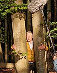 Eine Person in einem gelben Jackett steht zwischen zwei Bäumen und umschlingt sie mit den Armen. Im Hintergrund sind weitere Bäume und Laub sichtbar. Über dem Kopf der Person ist ein Mikrofon angebracht, das auf die Person gerichtet ist.
